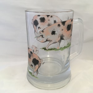 May include: Clear glass beer mug with a charming pig illustration. The pigs are painted in pink, black, and white, with green grass details. The mug has a handle and a solid base. Ideal for enjoying a pint of beer or other drinks.