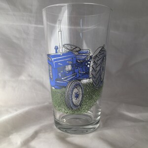 May include: A blue and white glass pint glass with a drawing of a blue tractor in a field of green grass.