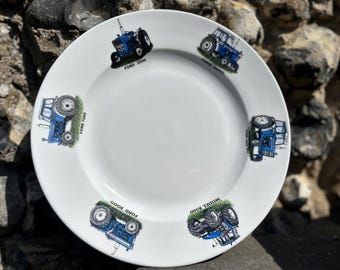 Könnte beinhalten: Ein weißer Keramikteller mit blauen und grünen Illustrationen von Ford-Traktoren. Der Teller zeigt vier verschiedene Modelle: Ford 3000, Ford 7000, Modell 8500 und Ford 3000.