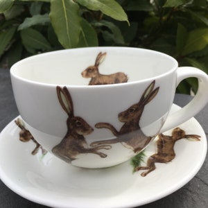 May include: White teacup and saucer set featuring brown hare illustrations. The teacup has a large handle and the saucer is round. The hares are depicted in various poses, adding a whimsical touch to the tea set.
