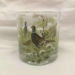 May include: A clear glass tumbler with a printed design of three pheasants in a forest setting. The pheasants are brown, green, and blue with white accents. The forest is depicted in shades of green and brown.