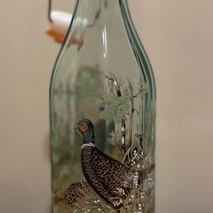 Peut inclure: Une bouteille en verre transparent avec une anse en fil de fer. La bouteille a un motif de faisan avec des couleurs brunes, noires et blanches.