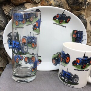 Puede incluir: Un juego de tres artículos de cocina de cerámica con un diseño de tractor azul y naranja. El juego incluye una taza, un vaso y un plato. La taza y el vaso tienen un patrón repetido de tractores, mientras que el plato tiene un solo diseño de tractor en el centro. El texto en los artículos dice "Fordson Dexta" y "Fordson Super Major".