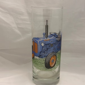 May include: A clear glass with a blue and orange tractor design. The tractor is on a green field.