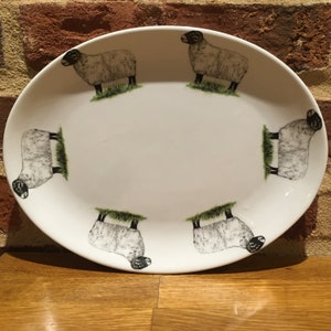 May include: White oval ceramic serving platter with a black and white sheep design. The sheep are standing on a green grass background.