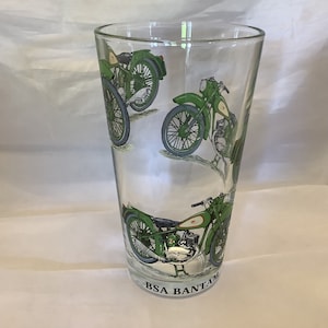 Pode incluir: Um copo de vidro transparente com um design verde e branco de motocicletas antigas. As motocicletas têm o texto "BSA Bantam" impresso na parte inferior do copo.
