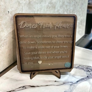 May include: A wooden plaque with a glass front, featuring the text "Dimes From Heaven". The plaque has a quote about finding dimes as a sign from an angel. The plaque is on a small wooden stand.