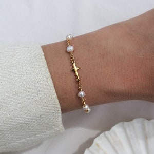 Peut inclure: Un bracelet en chaîne dorée avec un petit pendentif en forme de croix et des perles blanches.