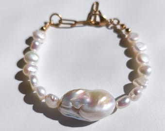 Puede incluir: Una delicada pulsera de perlas con una gran perla barroca iridiscente como pieza central. La pulsera está ensartada con perlas más pequeñas de forma irregular y conectada con un cierre dorado. La joya está sobre un fondo blanco.
