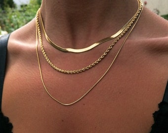 May include: Three gold chain necklaces layered on a person's neck. The chains are different styles and thicknesses.