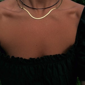 Puede incluir: Un collar de cadena dorada superpuesto a un collar de cuentas negras. Ambos collares se llevan en el cuello de una persona.