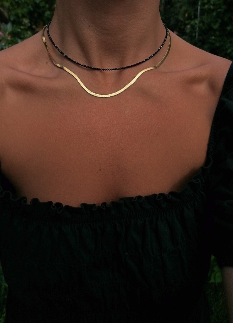 Puede incluir: Un collar de cadena dorada superpuesto a un collar de cuentas negras. Ambos collares se llevan en el cuello de una persona.