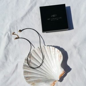 Puede incluir: Un collar de cuentas negras con cierres dorados, presentado sobre una concha blanca, con una caja negra con la etiqueta "By Isabelle Fashion" en el fondo.