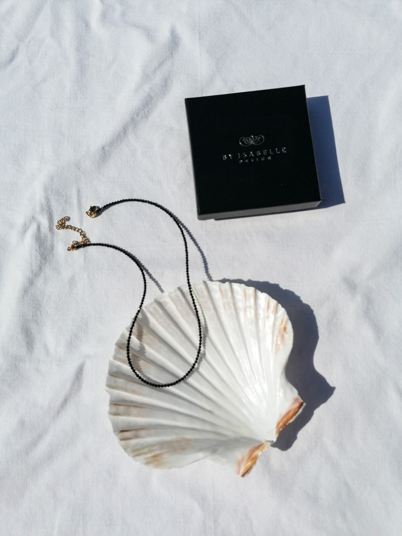 Puede incluir: Un collar de cuentas negras con cierres dorados, presentado sobre una concha blanca, con una caja negra con la etiqueta "By Isabelle Fashion" en el fondo.
