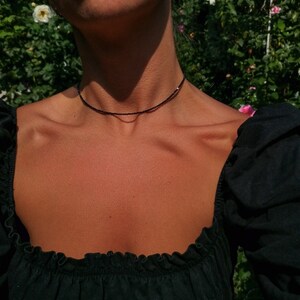 Puede incluir: Una mujer con un top negro de hombros descubiertos y un delicado collar negro de gargantilla. El collar est&aacute; hecho de peque&ntilde;as cuentas negras.