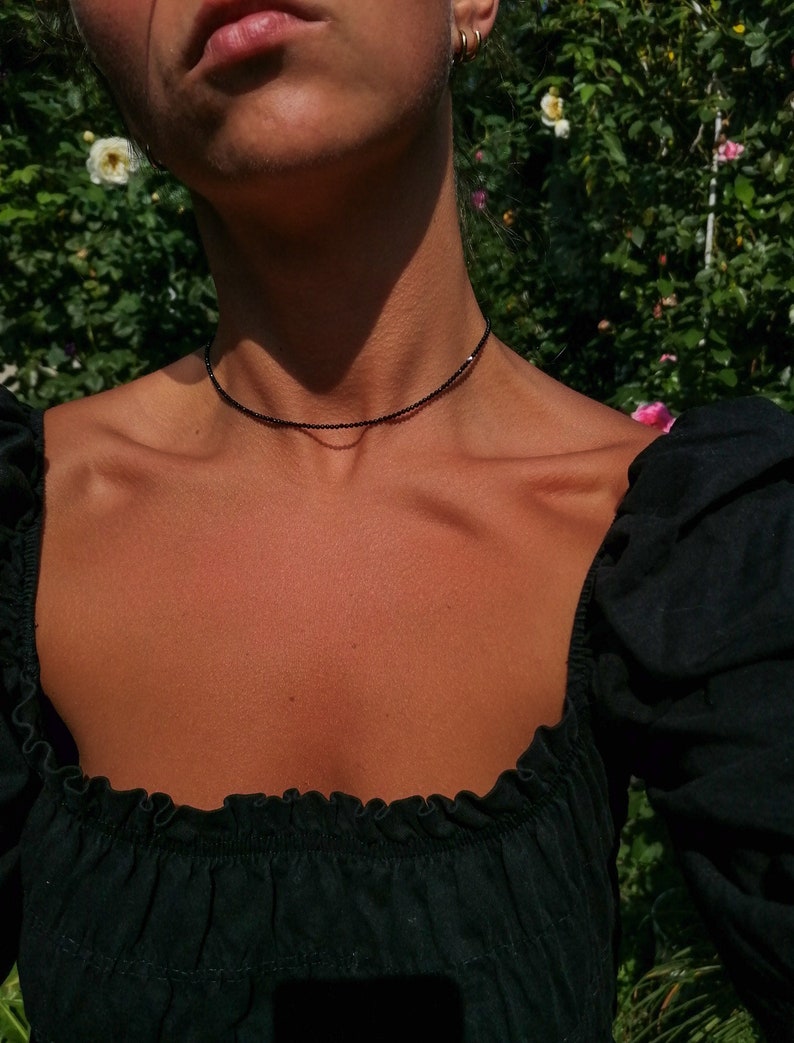 Puede incluir: Una mujer con un top negro de hombros descubiertos y un delicado collar negro de gargantilla. El collar est&aacute; hecho de peque&ntilde;as cuentas negras.