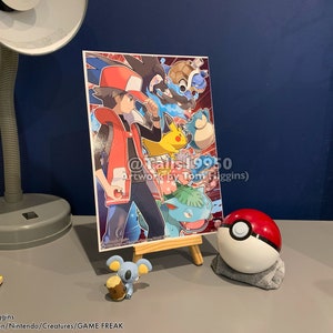 K&ouml;nnte beinhalten: Ein roter und wei&szlig;er Pok&eacute;ball mit einem schwarzen Streifen, eine kleine graue und braune Pok&eacute;mon-Figur und ein gerahmter Druck eines Comic-Charakters mit roten Haaren und einer rot-wei&szlig;en Weste, umgeben von Pok&eacute;mon-Charakteren. Der Druck enth&auml;lt den Text "@Talis19950 Artwork by Tom Higgins)"