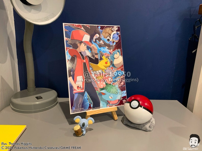 K&ouml;nnte beinhalten: Ein roter und wei&szlig;er Pok&eacute;ball mit einem schwarzen Streifen, eine kleine graue und braune Pok&eacute;mon-Figur und ein gerahmter Druck eines Comic-Charakters mit roten Haaren und einer rot-wei&szlig;en Weste, umgeben von Pok&eacute;mon-Charakteren. Der Druck enth&auml;lt den Text "@Talis19950 Artwork by Tom Higgins)"