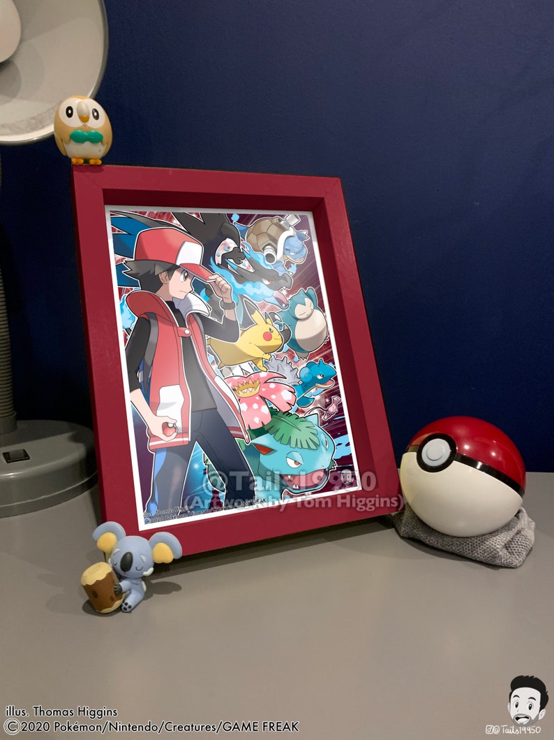 K&ouml;nnte beinhalten: Ein gerahmter Druck einer Pok&eacute;mon-Illustration mit einem rothaarigen Trainer mit einem Pikachu und anderen Pok&eacute;mon. Der Druck ist in einem roten Rahmen und hat den Text "Artwork by Tom Higgins" unten.