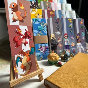 Może przedstawiać: Zestaw kolorowych zakładek do książek z postaciami Pokemonów, przedstawiającymi Charmandera, Charmeleona i Charizarda. Zakładki są wystawione na drewnianym stojaku i otoczone innymi przedmiotami z motywem Pokemonów.