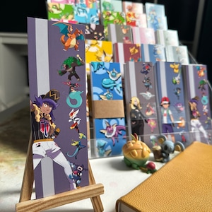 Puede incluir: Un marcapáginas morado y blanco con un personaje de la serie de anime Pokémon. El marcapáginas presenta una variedad de personajes Pokémon, incluyendo Charizard, Snorlax y Pikachu. El marcapáginas se muestra en un pequeño caballete de madera.