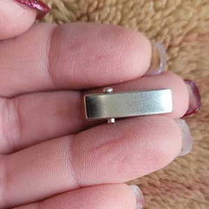 May include: A silver-toned metal clasp for a bracelet or necklace. The clasp is rectangular in shape and has a smooth, polished finish.