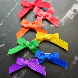 May include: Seven rainbow-colored hair bows arranged in a rainbow order from top to bottom. The bows are made of satin ribbon and are tied in a simple bow shape.