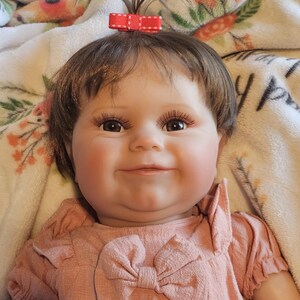 May include: A smiling baby doll with brown hair, wearing a pink dress with a bow. The doll has a red bow in her hair and is lying on a white blanket with floral designs.