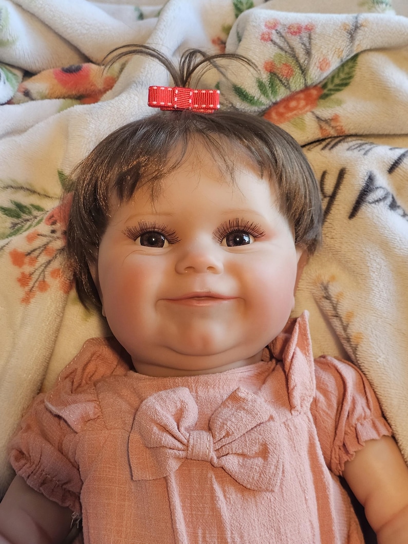 May include: A smiling baby doll with brown hair, wearing a pink dress with a bow. The doll has a red bow in her hair and is lying on a white blanket with floral designs.