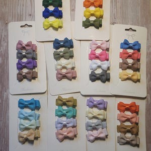 May include: Sets of colorful bow hair clips in various shades, including red, blue, green, and yellow. Each set contains three bows attached to a white card. The bows are made of a ribbed fabric.