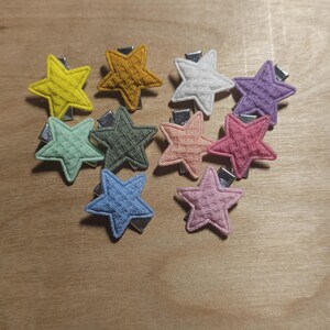 May include: A collection of ten star-shaped hair clips in various colors, including yellow, gold, white, purple, green, peach, pink, and blue. Each clip has a textured fabric design and a silver metal clip.