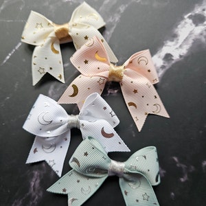 May include: Four small hair bows with a moon and star pattern. The bows are in white, pink, and green.