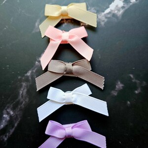 May include: Five small satin ribbon bows in different colors. The bows are arranged vertically from top to bottom in the following order: pale yellow, light pink, brown, white, and light purple.