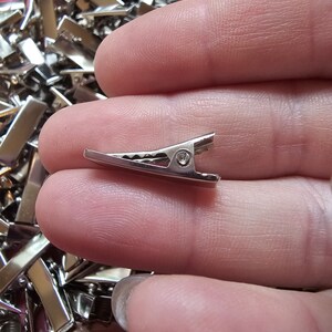May include: A close-up of a silver metal alligator clip. The clip is small and has a spring-loaded mechanism.