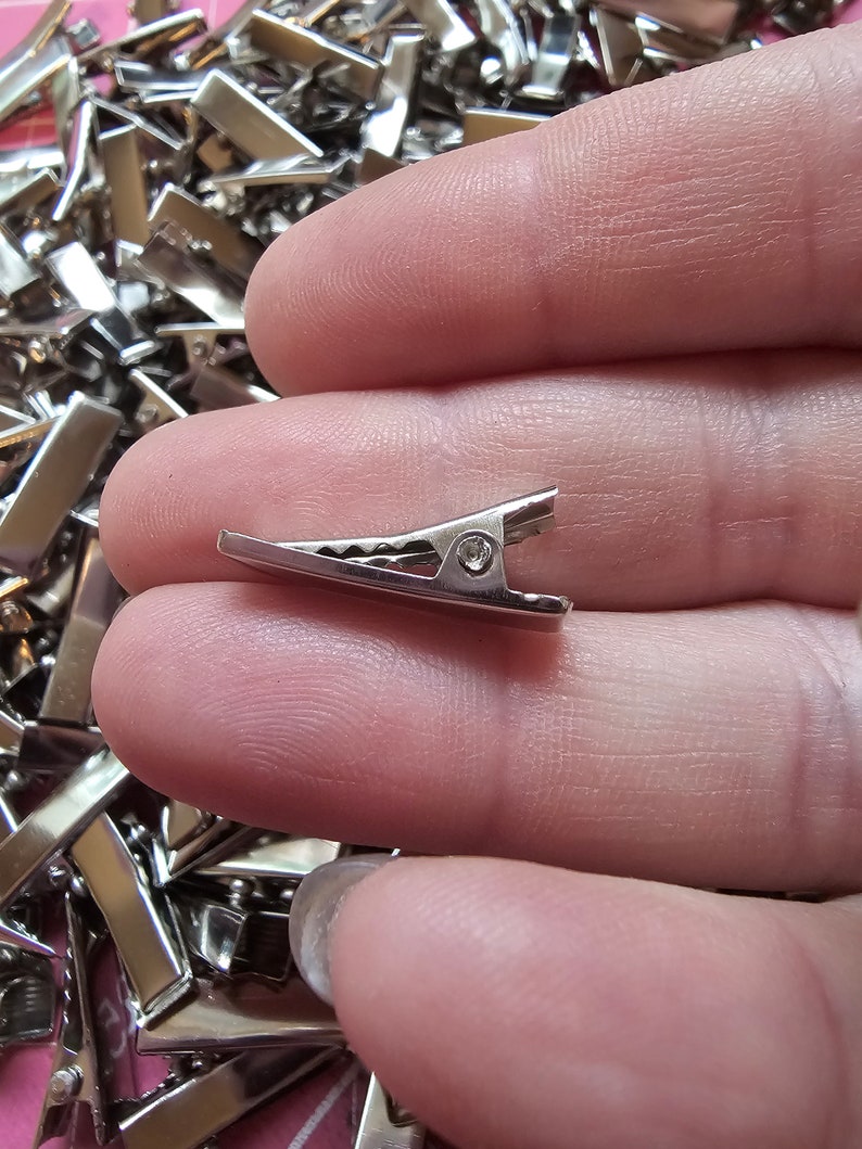 May include: A close-up of a silver metal alligator clip. The clip is small and has a spring-loaded mechanism.