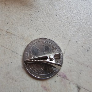 May include: A silver-colored metal binder clip is placed on top of a 2015 North Carolina quarter. The quarter features a profile of the Wright brothers and their airplane.