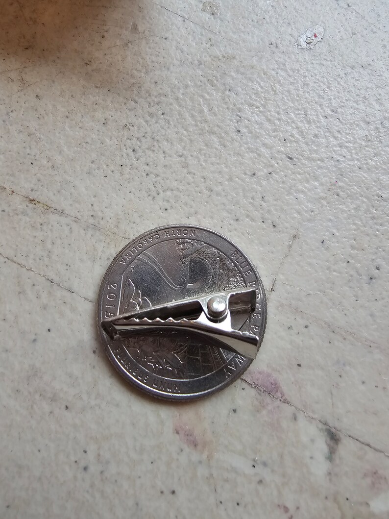 May include: A silver-colored metal binder clip is placed on top of a 2015 North Carolina quarter. The quarter features a profile of the Wright brothers and their airplane.