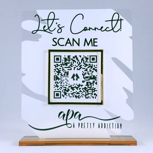 May include: White acrylic sign with black text that reads "Let's Connect! SCAN ME". A black and white QR code is in a gold frame. The text "apa A PRETTY ADDICTION" is below the QR code.