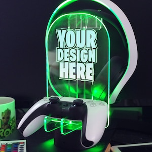 May include: A white and black video game controller stand with a green LED light. The stand has a slot for a headset and a space for a controller. The stand has a clear acrylic panel with the text "YOUR DESIGN HERE" on it.