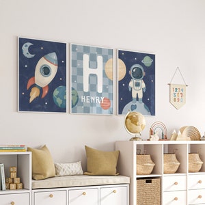 May include: A white bookshelf with four shelves and two drawers, a white bench with four drawers, and a white bookshelf with six shelves. The shelves are filled with books, toys, and baskets. The walls are white and there are three framed prints of an astronaut, a rocket ship, and the letter H with the name Henry. There is a wooden sign that says "Explore" on the wall.