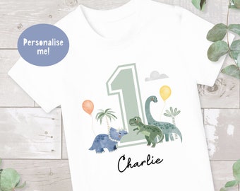 May include: White t-shirt with a green number one, a green dinosaur, a blue dinosaur, and the name "Charlie" printed on it. The shirt is decorated with a green palm tree, a yellow balloon, and a pink balloon.