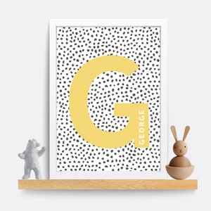 Puede incluir: Una impresi&oacute;n enmarcada en blanco con un fondo de lunares blanco y negro. Una gran letra amarilla "G" est&aacute; en el centro de la impresi&oacute;n con el nombre "GEORGE" escrito verticalmente a lo largo del lado derecho de la letra.