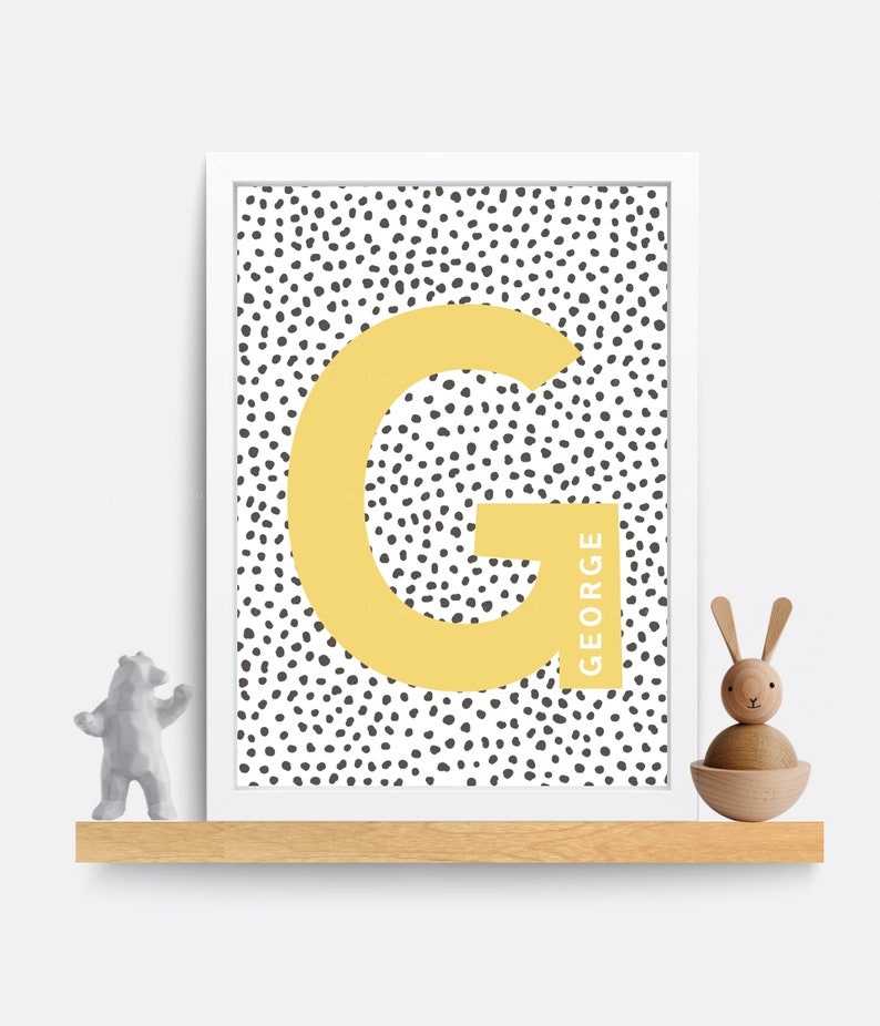 Puede incluir: Una impresi&oacute;n enmarcada en blanco con un fondo de lunares blanco y negro. Una gran letra amarilla "G" est&aacute; en el centro de la impresi&oacute;n con el nombre "GEORGE" escrito verticalmente a lo largo del lado derecho de la letra.