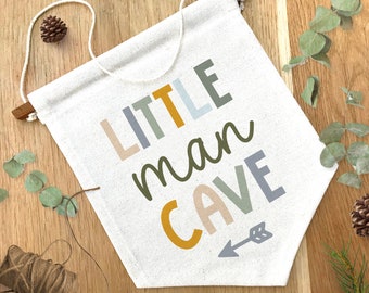 May include: A white canvas banner with the text "LITTLE man CAVE" in colorful letters. The letters are in different shades of yellow, green, blue, and gray. An arrow is drawn in gray below the text.