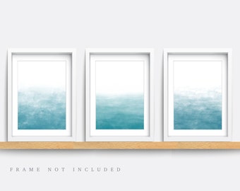 May include: Set of three framed watercolor prints with a gradient of blue shades, resembling a sky or ocean scene. The frames are white and the prints are displayed on a wooden shelf. "FRAME NOT INCLUDED" is written below the shelf.