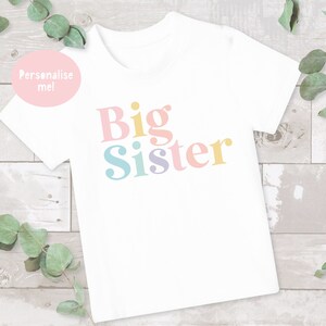 May include: White t-shirt with the text "Big Sister" in colorful letters. The letters are in a retro font style.