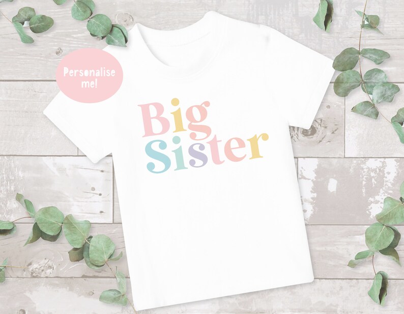 May include: White t-shirt with the text "Big Sister" in colorful letters. The letters are in a retro font style.