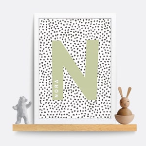 Puede incluir: Una impresi&oacute;n enmarcada en blanco con una letra N verde sobre un fondo blanco con lunares negros. El nombre "NORA" est&aacute; escrito verticalmente en el lado izquierdo de la letra N.