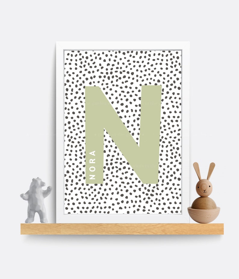 Puede incluir: Una impresi&oacute;n enmarcada en blanco con una letra N verde sobre un fondo blanco con lunares negros. El nombre "NORA" est&aacute; escrito verticalmente en el lado izquierdo de la letra N.