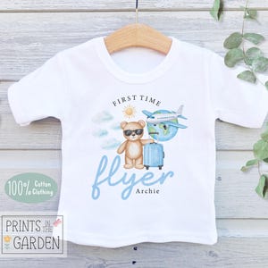 May include: White cotton t-shirt featuring a teddy bear wearing sunglasses, holding a blue suitcase, and standing next to a globe and airplane. The shirt has the text "First Time Flyer" and "Archie" in blue, with a sun and clouds design.